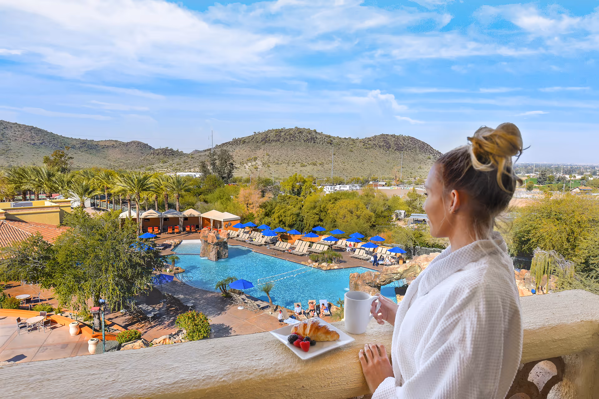 Woman in white robe holding a coffee cup on a balcony overlooking a resort pool with blue umbrellas and surrounding desert hills.